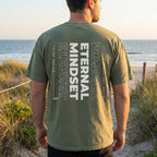Person wearing a green t-shirt with text on the back, standing on a beach.