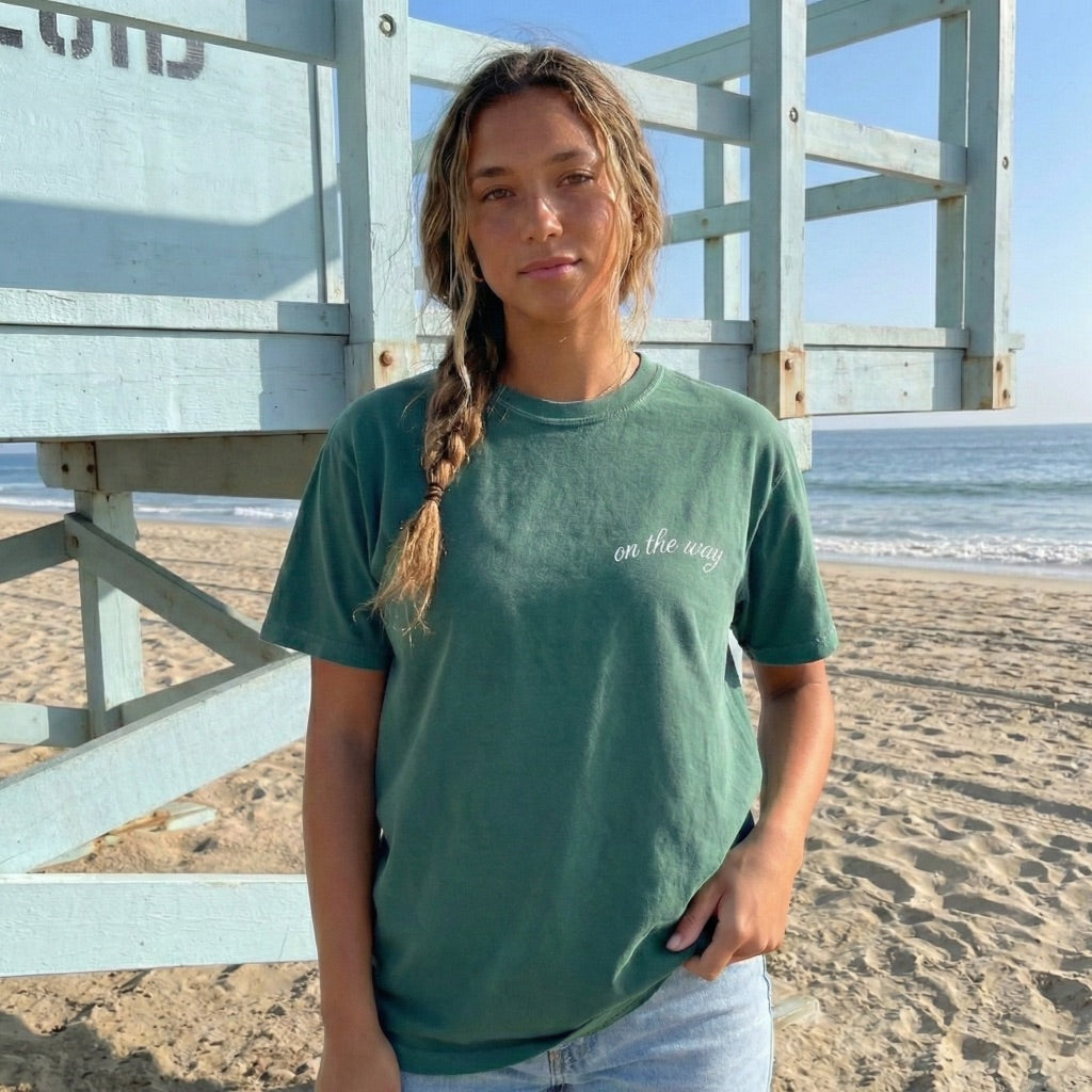 Back view of woman in teal On The Way t-shirt with minimalist mountain road graphic at the beach